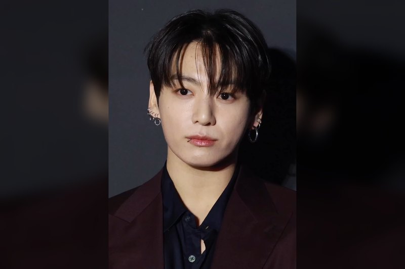 Close-up of Jung Kook with a dark fringe, a lip ring and silver hoop earrings, wearing a burgundy suit jacket over a bla
