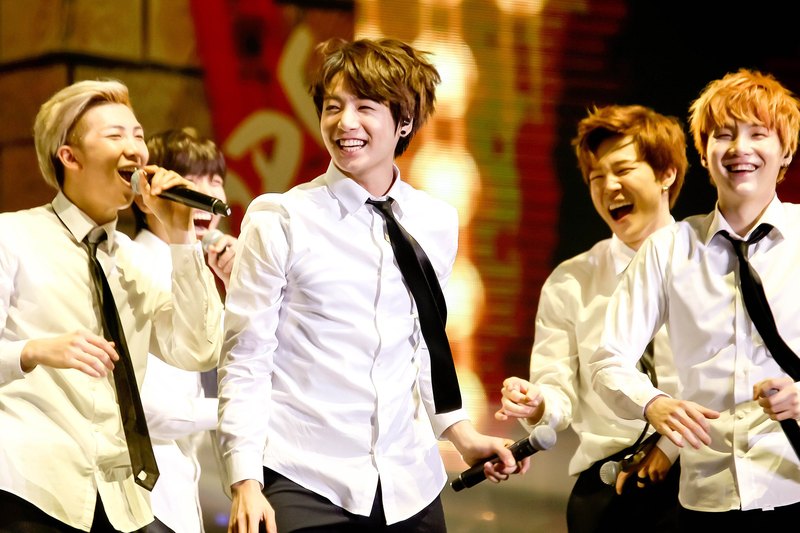 Five members in white shirts and black ties laughing and singing into microphones in front of a glowing stage screen