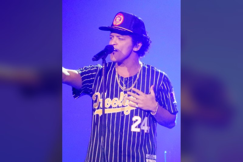 Bruno Mars singing into a microphone on stage under blue light, wearing a cap, gold chains and a pinstriped baseball jer
