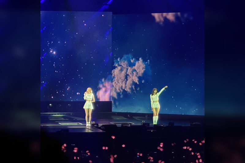 Two performers in white outfits on a large stage in front of a starry blue video screen, with pink light sticks glowing 